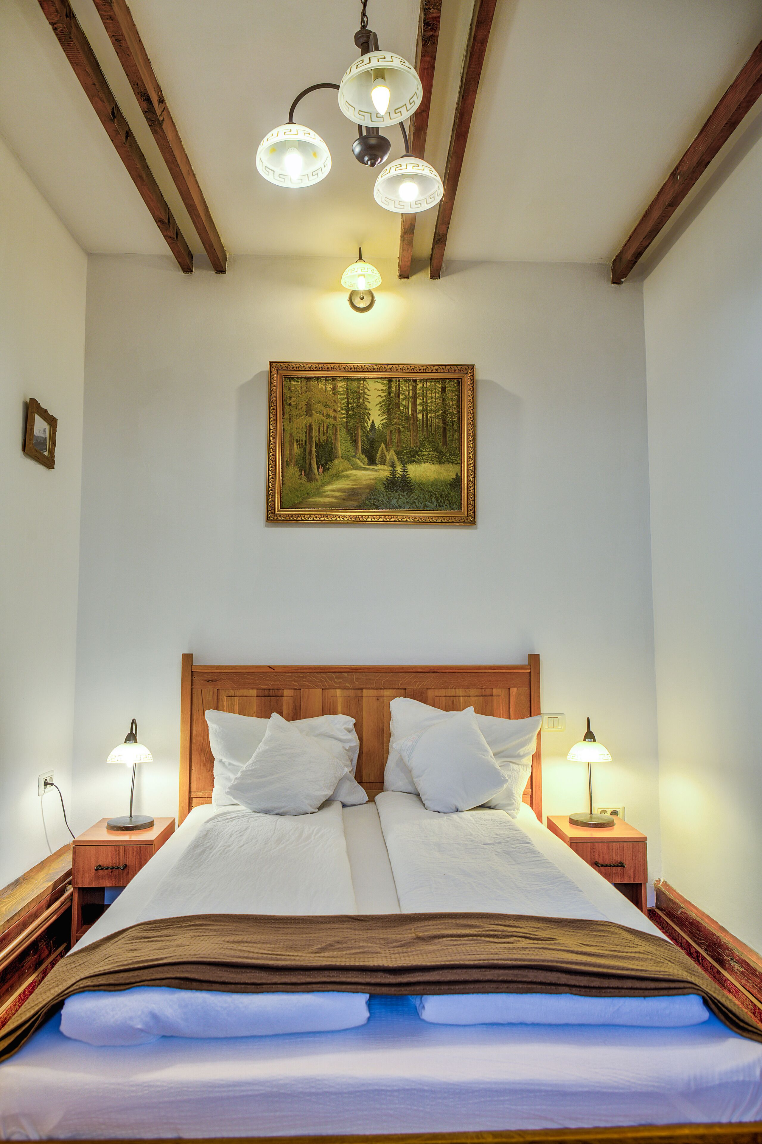 A neatly made bed with a wooden headboard, two bedside tables with lamps, and a forest-themed painting above the bed.