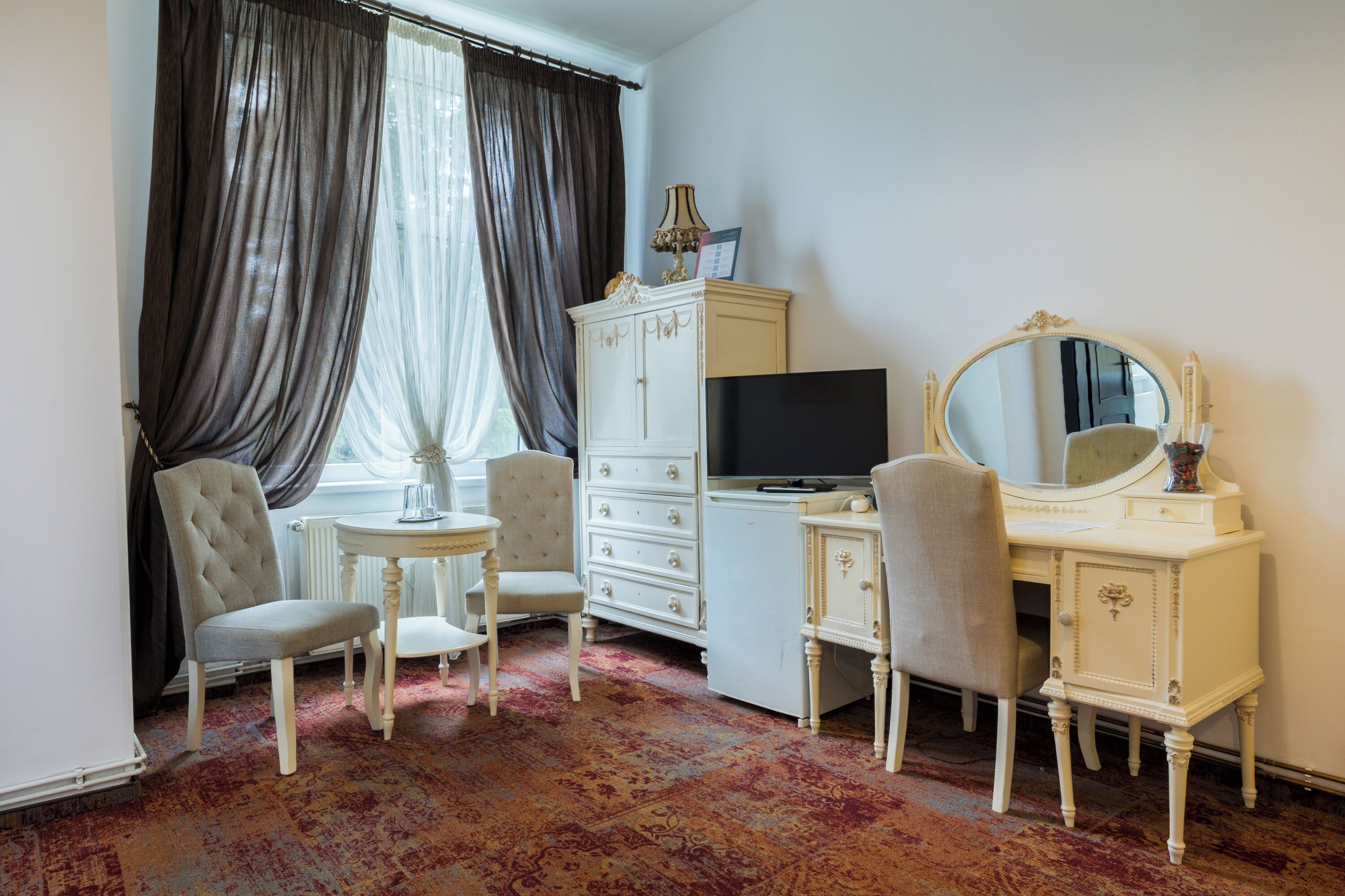 A room with a white vanity, a large mirror, a television, and two chairs.