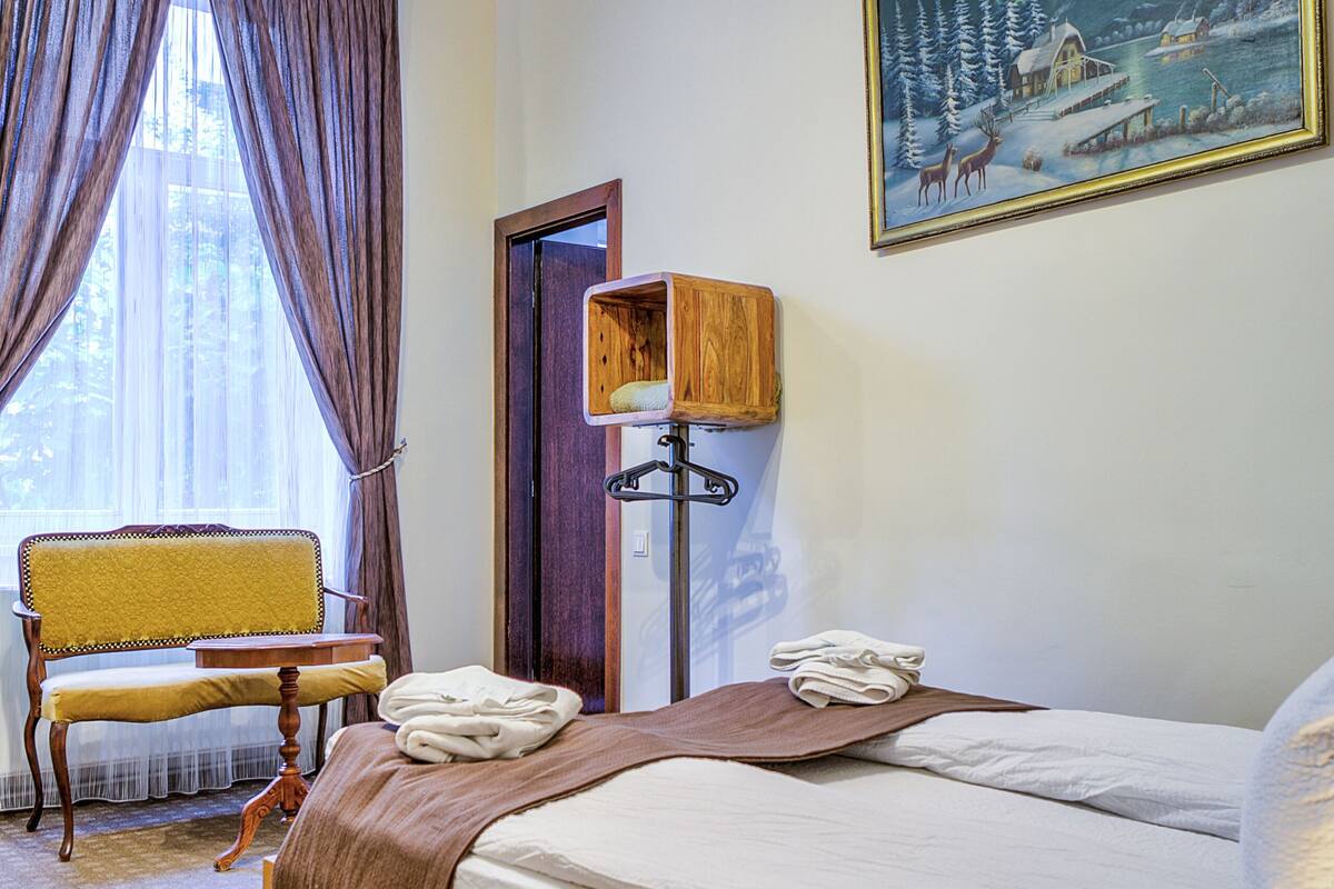 Quarto de hotel com uma cama grande, uma cadeira de madeira, escrivaninha, mesa de cabeceira, um quadro e um candelabro.
