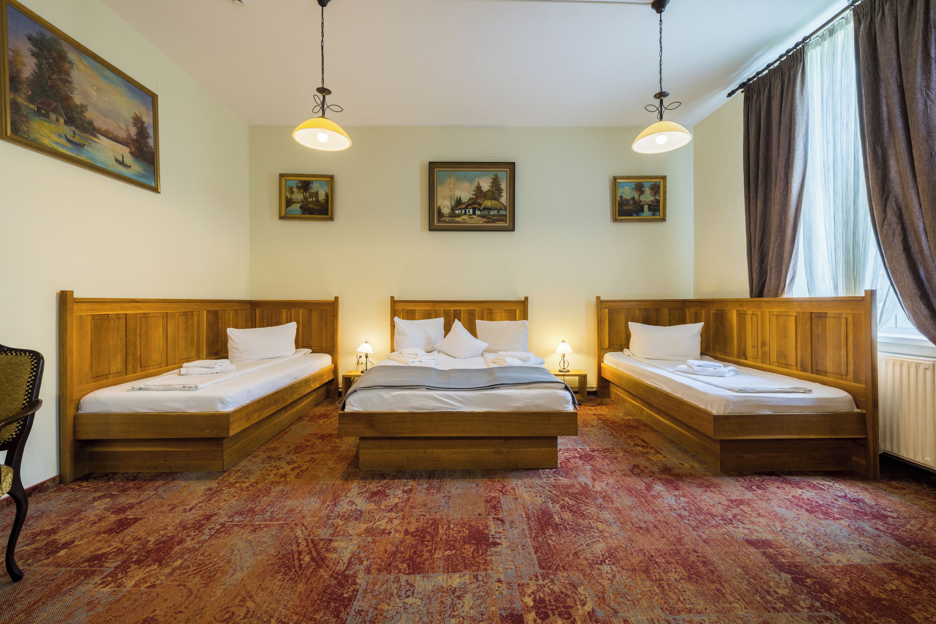 A hotel room with three wooden beds, a large window with curtains, and wall-mounted paintings.