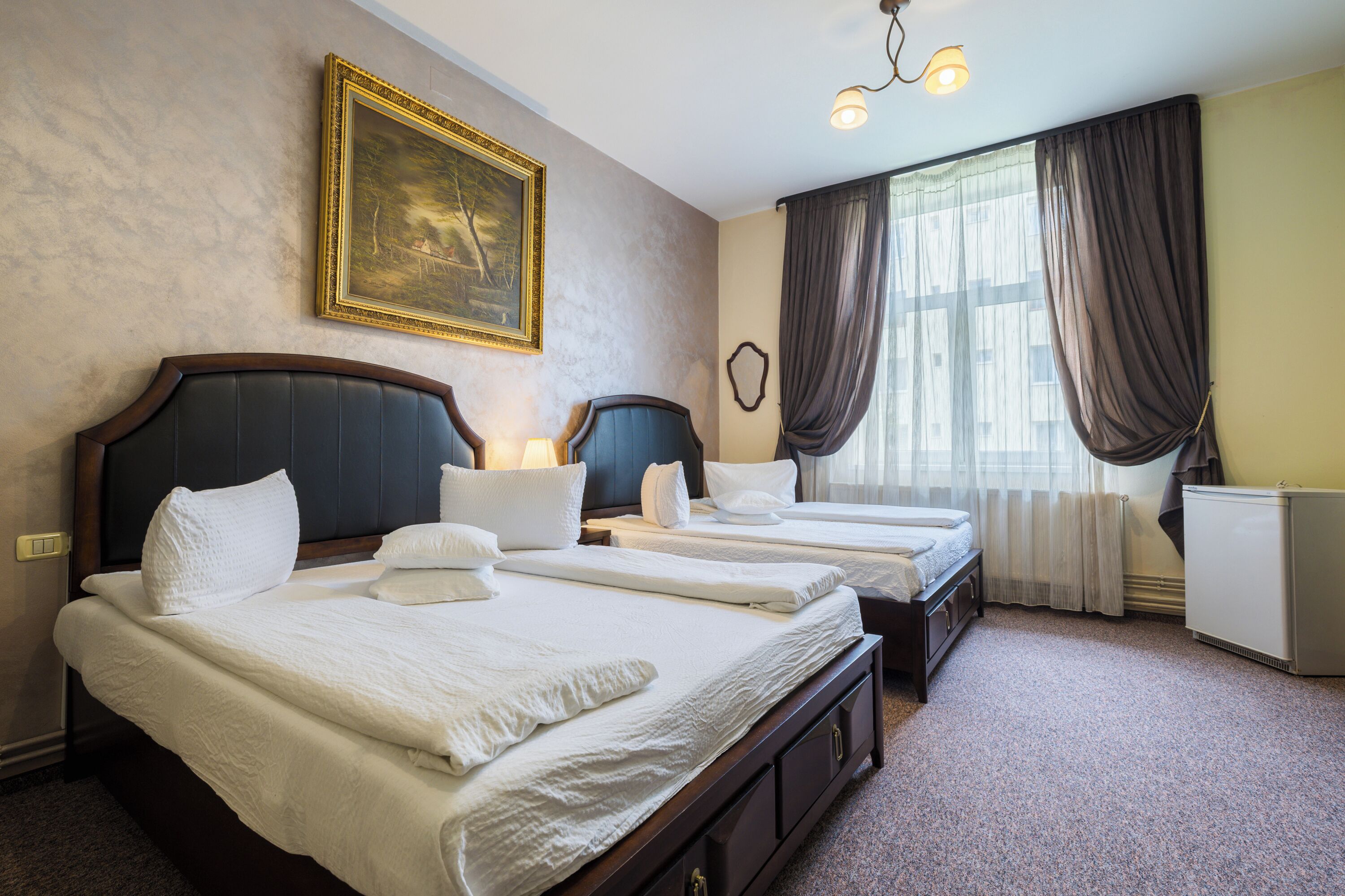 Une chambre d’hôtel avec trois lits, un tableau au mur, une fenêtre avec des rideaux et un radiateur.