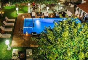 Una piscina al aire libre con sillones de descanso y mesas, rodeada por una terraza de madera y un césped cuidado.