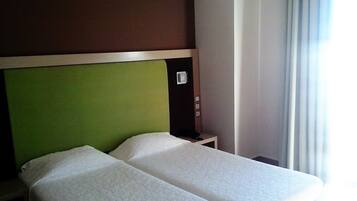 Habitación de hotel con cama doble, cabecera verde, mesita de noche pequeña y ventana con cortinas.