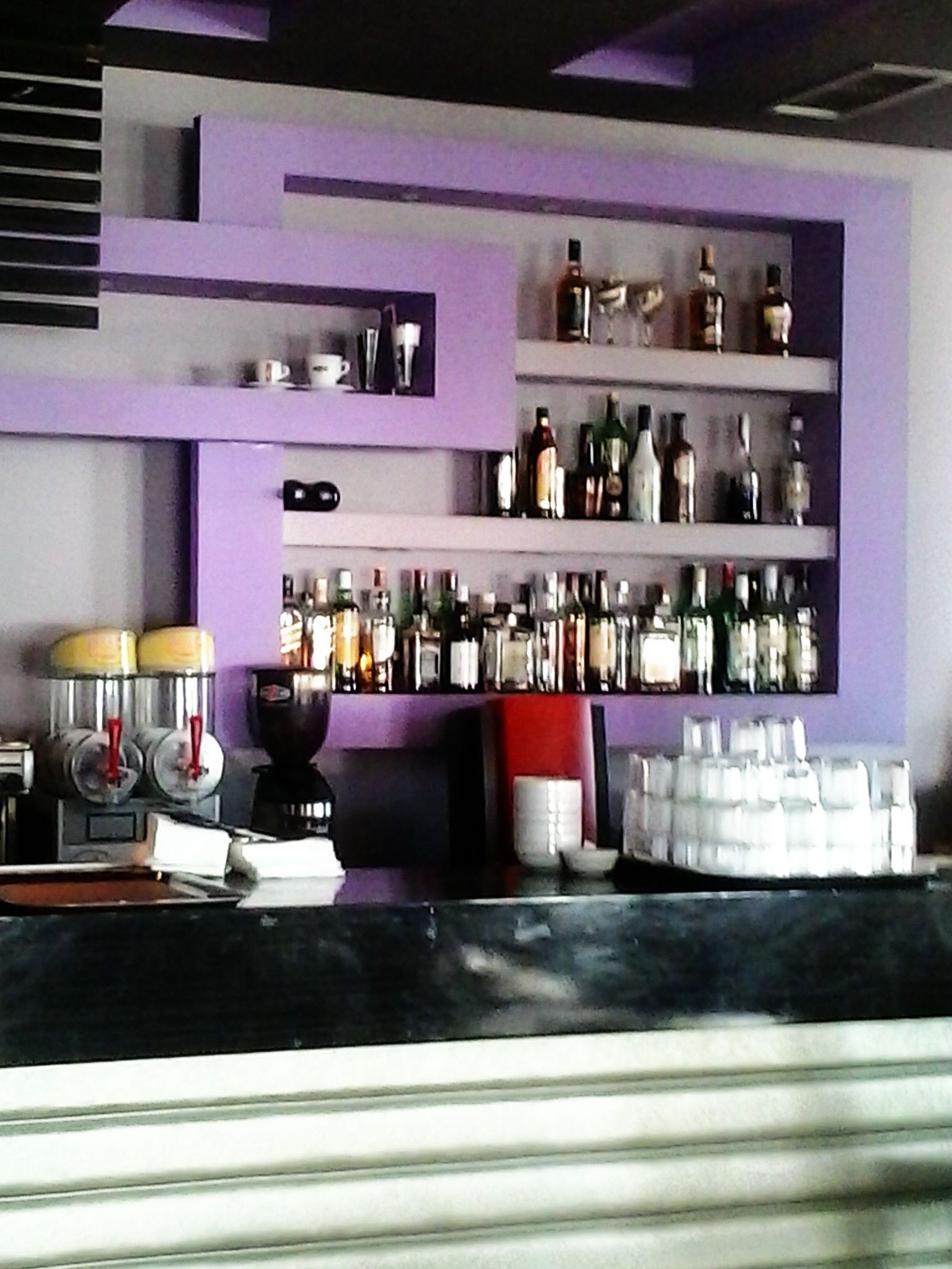 A bar area with shelves of bottles and glasses, a coffee machine, and a counter with a cash register.