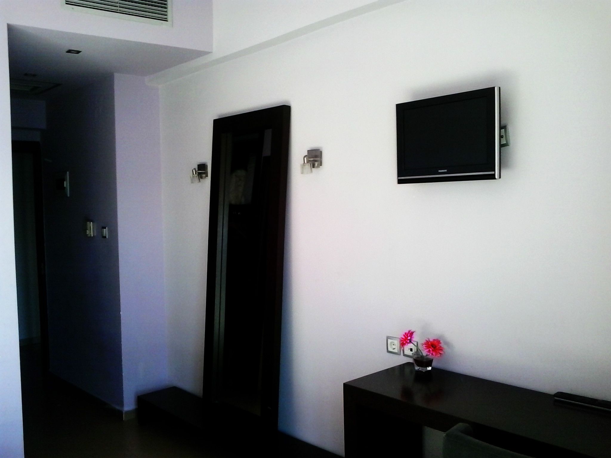 Ein Hotelzimmer mit flachbildfernseher an der Wand, einem Schreibtisch mit Blumenvase und einer Tür zu einem weiteren Zimmer.