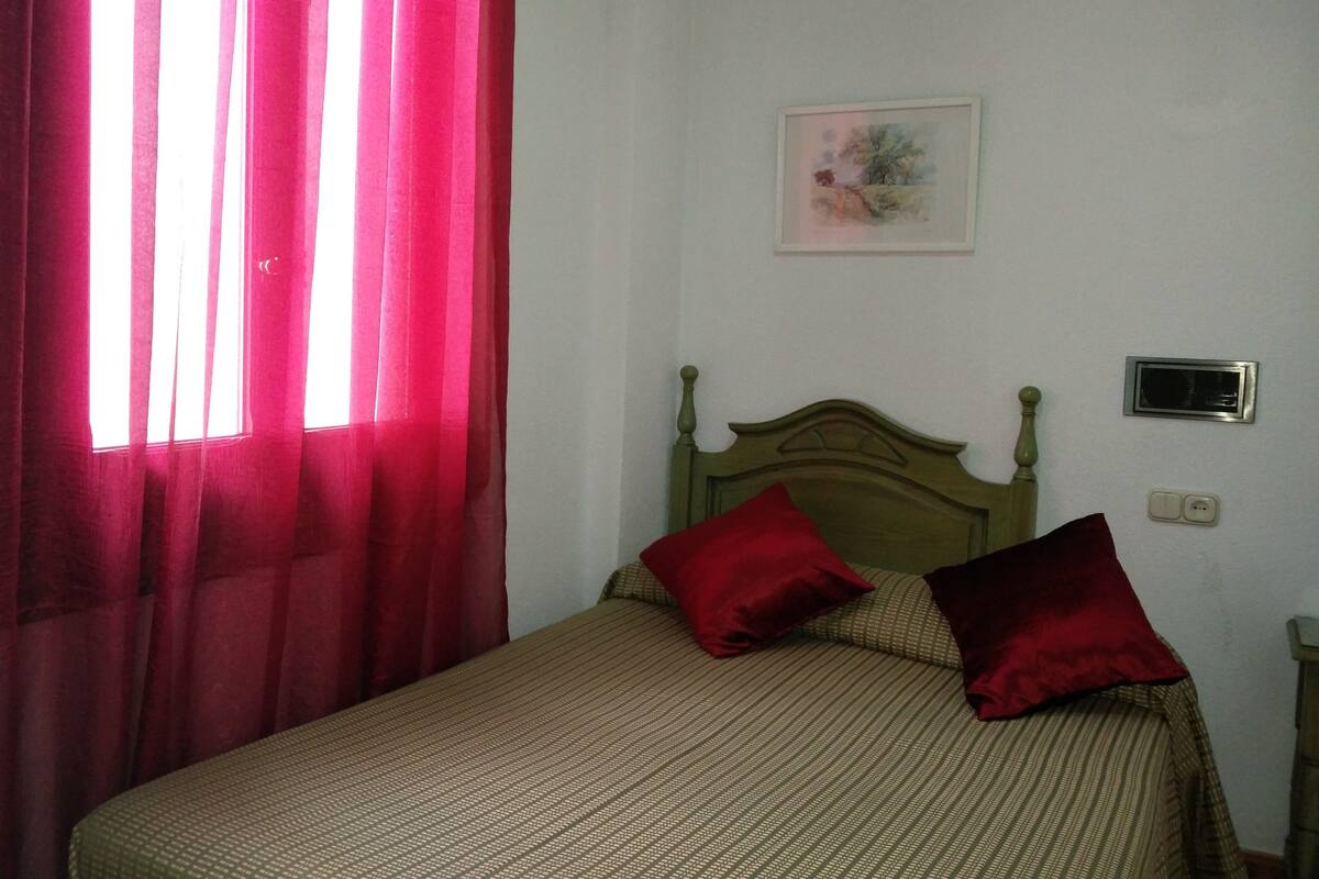 Una cama con cabecera de madera, almohadas rojas y una colcha verde en una habitación con paredes blancas y una ventana con cortinas rojas.