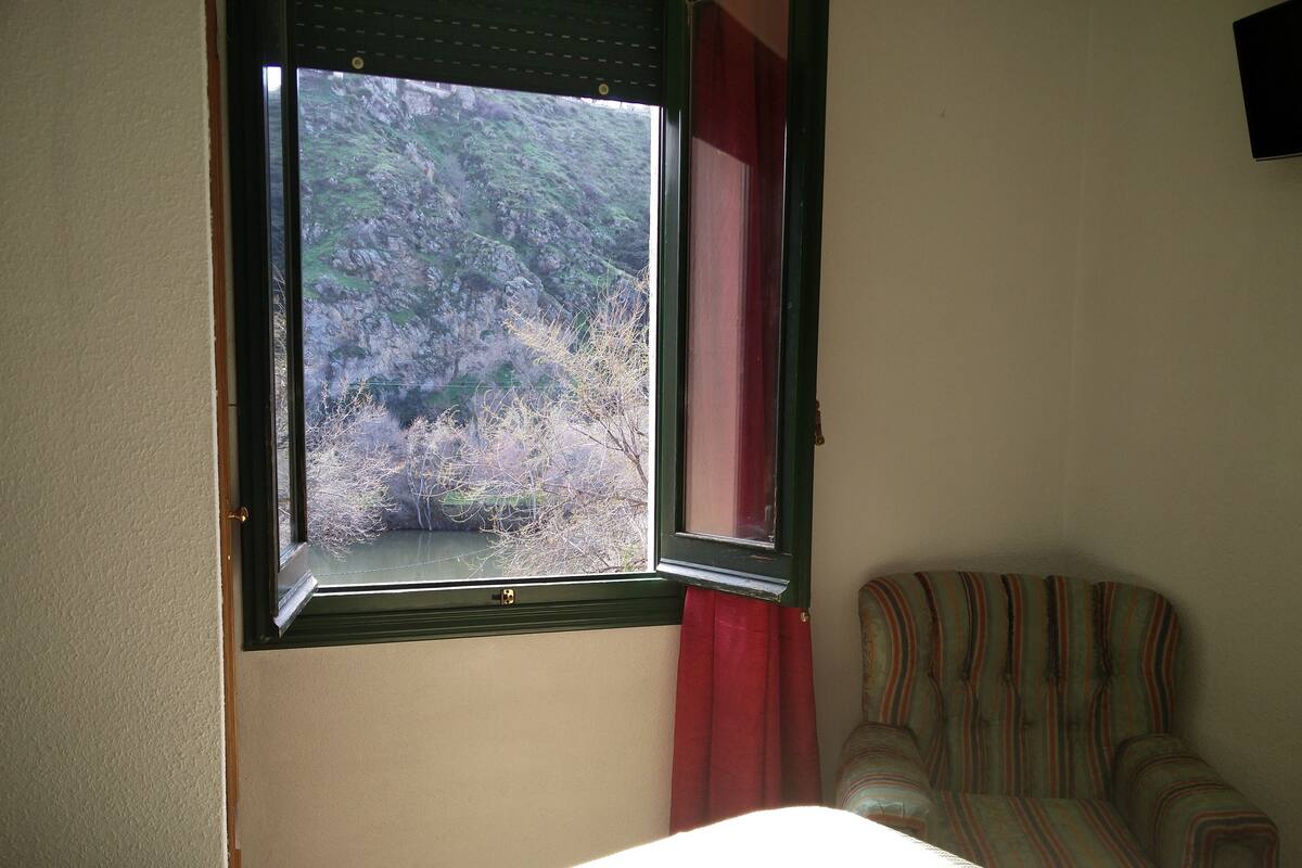 Una habitación con una cama, una ventana que da a árboles y una silla a rayas.