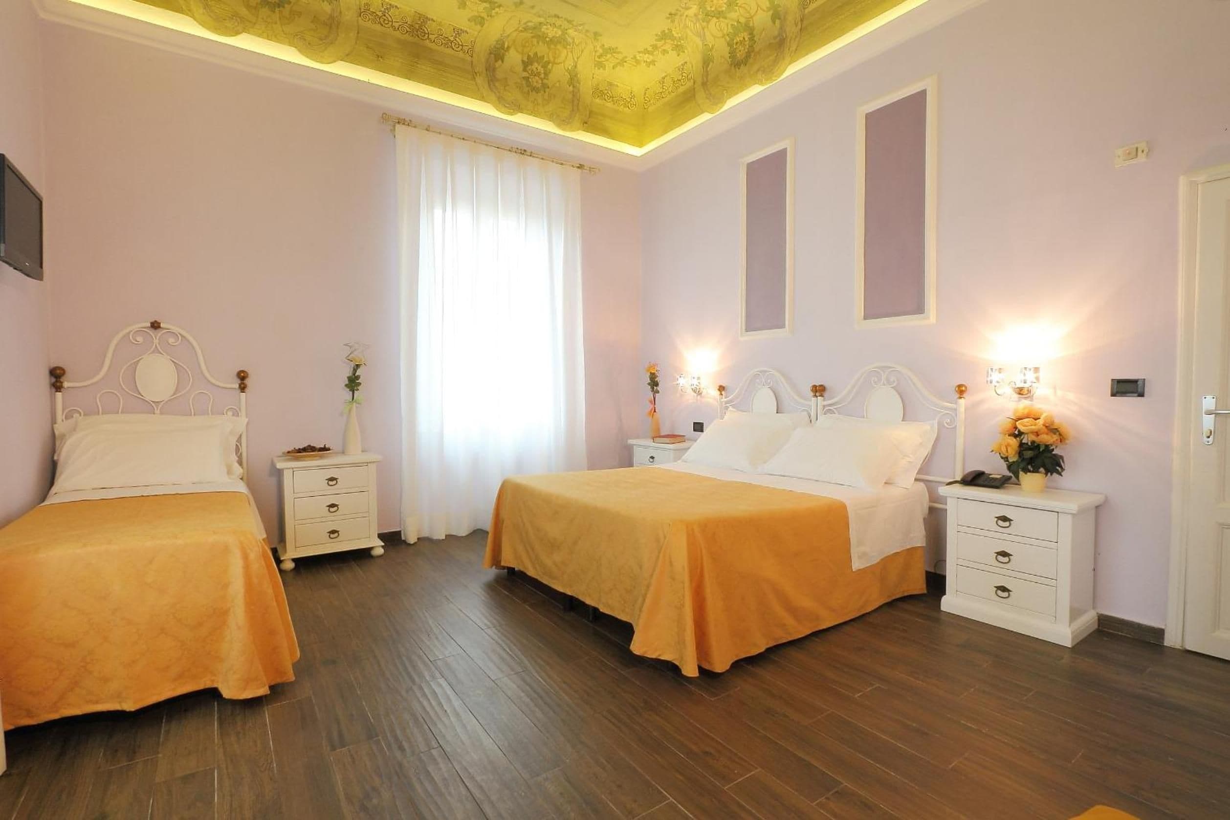Habitación de hotel con dos camas, piso de madera, techo con estampado floral y ventana con cortinas blancas.