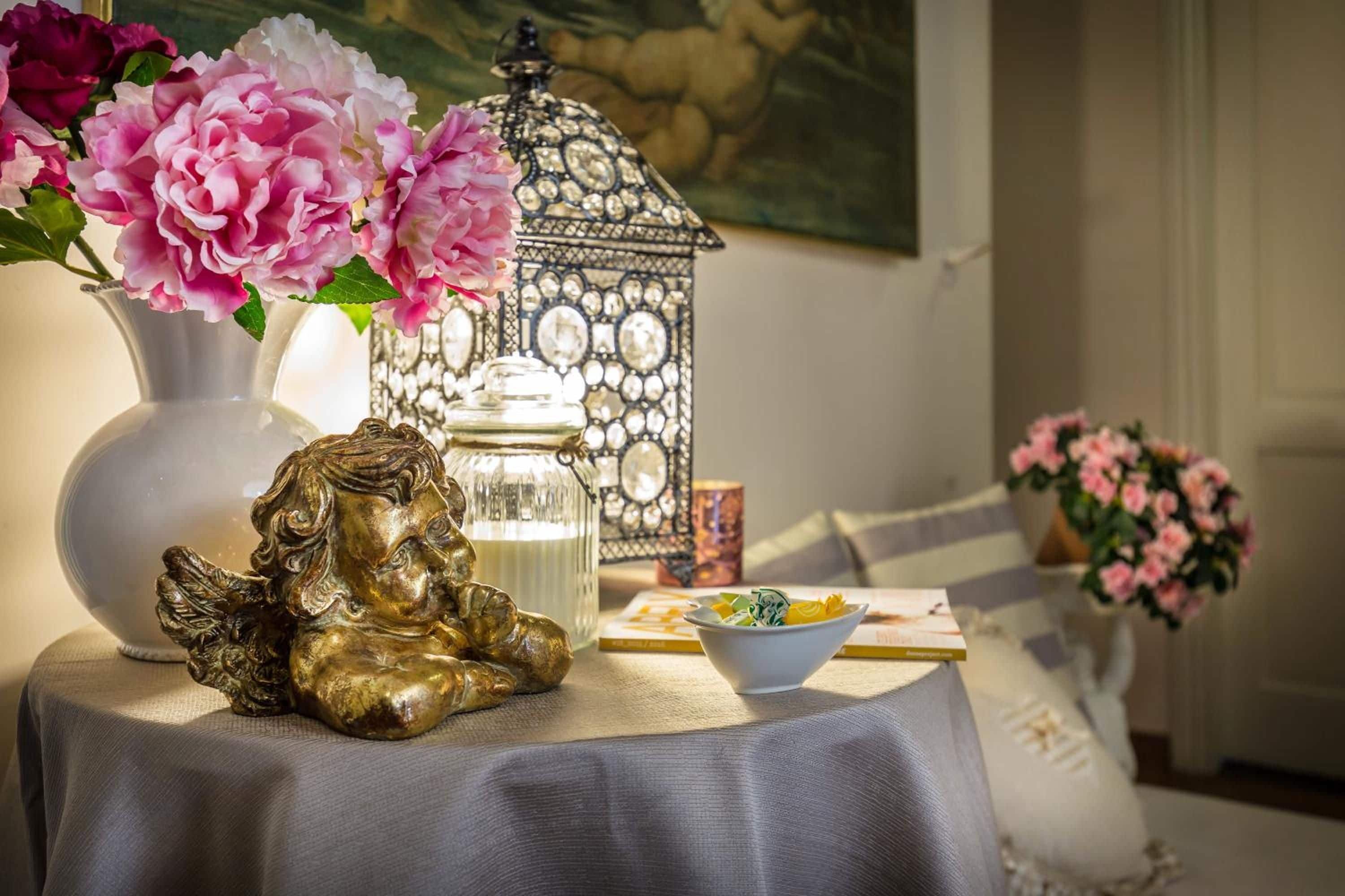 Una mesa con una figurita decorativa de un ángel, un florero con flores rosas, un farolillo y un cuenco de frutas.