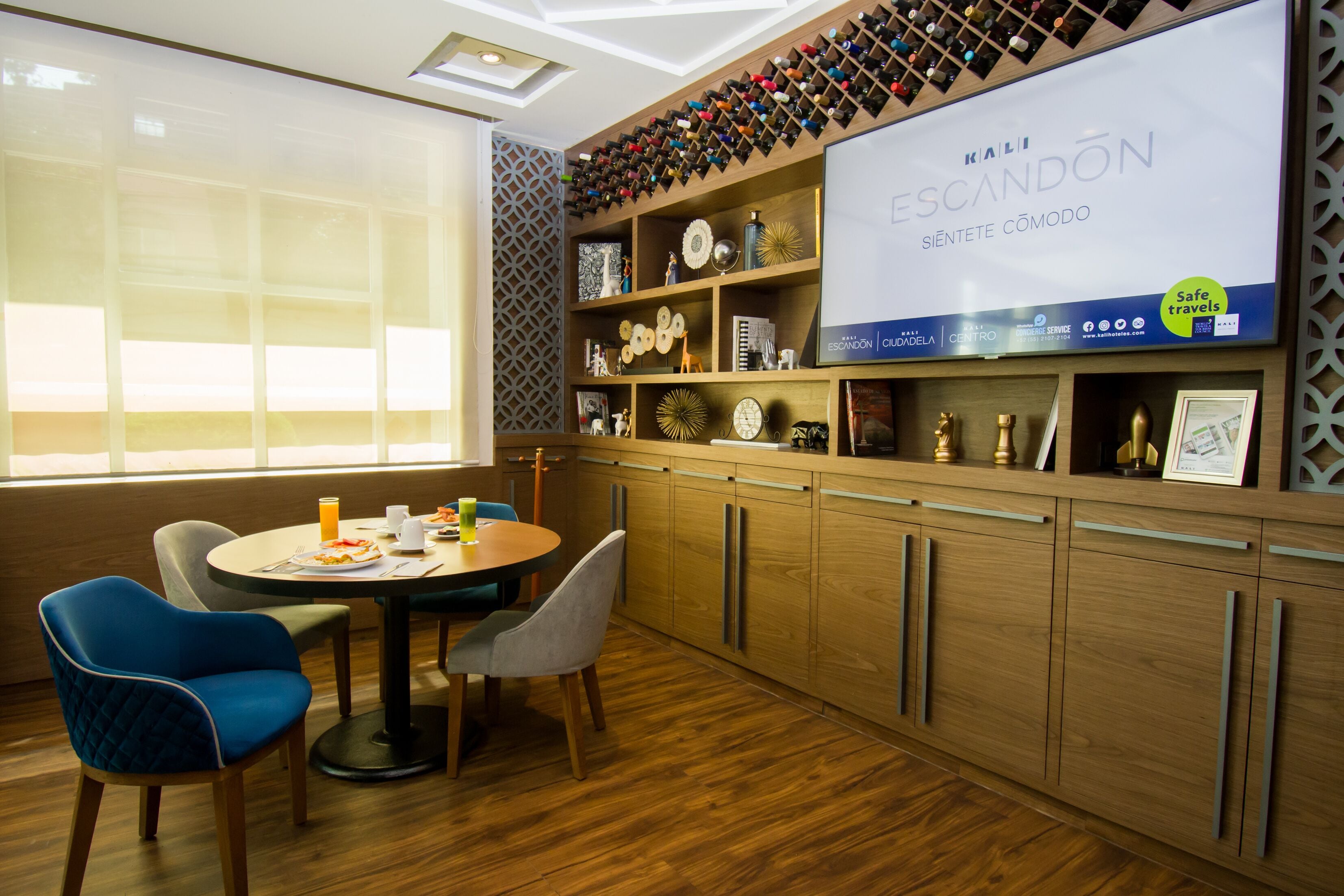 Habitación de hotel moderna con piso de madera, un área de comedor con una mesa redonda y sillas, y una repisa de pared con diversos objetos decorativos.