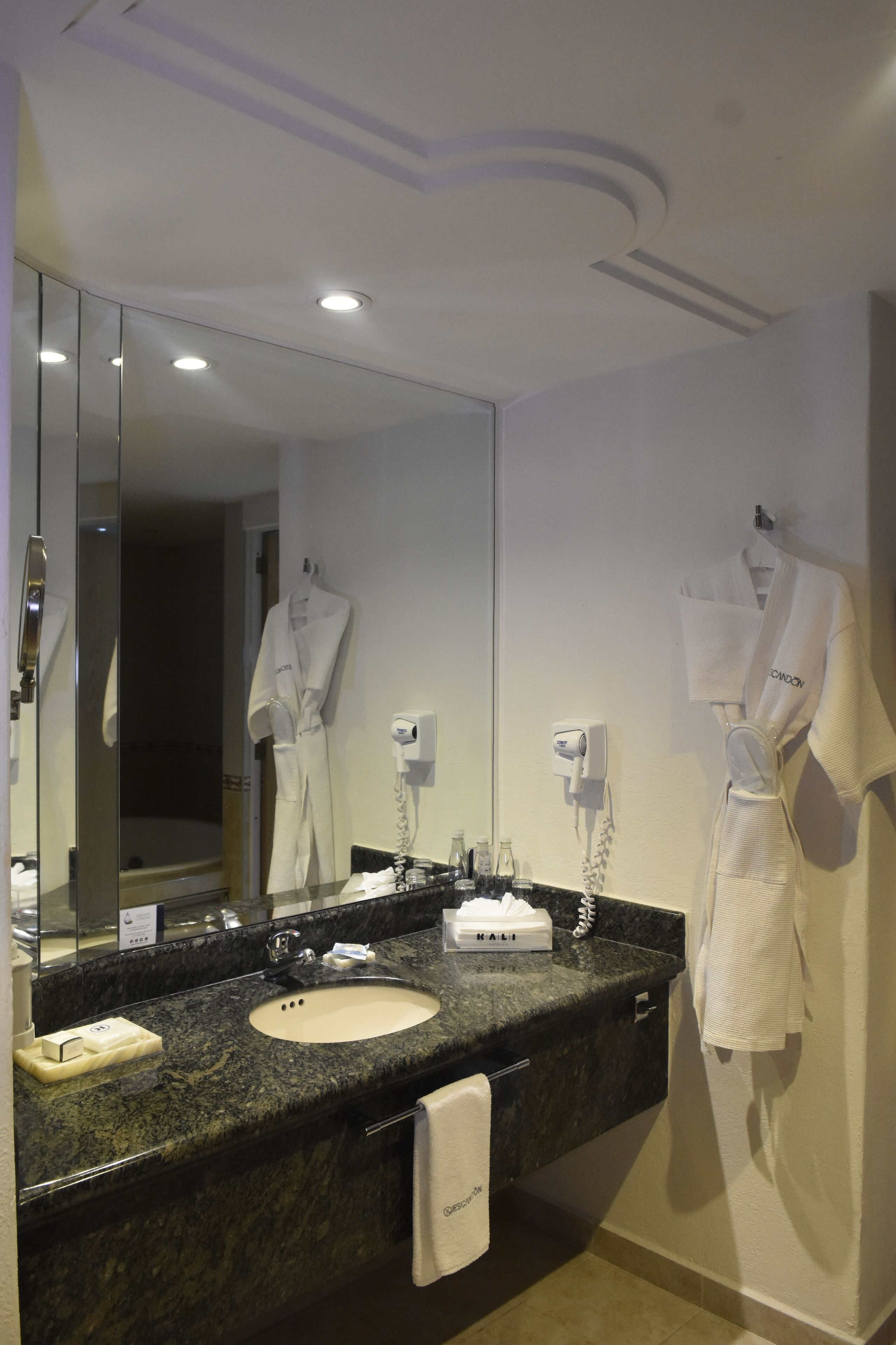 Un baño moderno con encimera de granito, un lavabo, un espejo y dos albornoces blancos colgados en la pared.