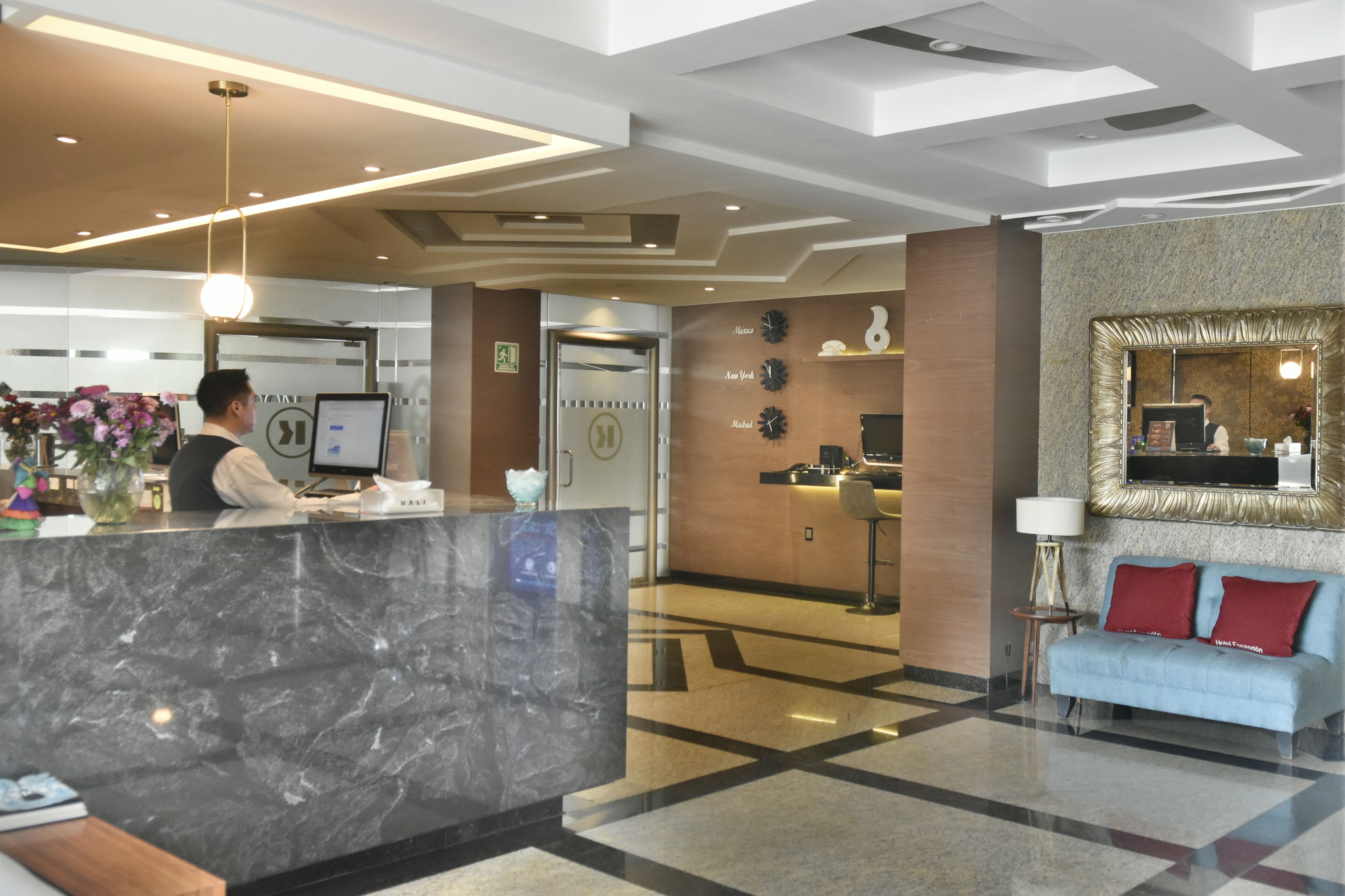 Zona de recepción de hotel con mostrador de mármol, una recepcionista, un escritorio de recepción, un sofá con cojines rojos y una pared con relojes y un espejo.
