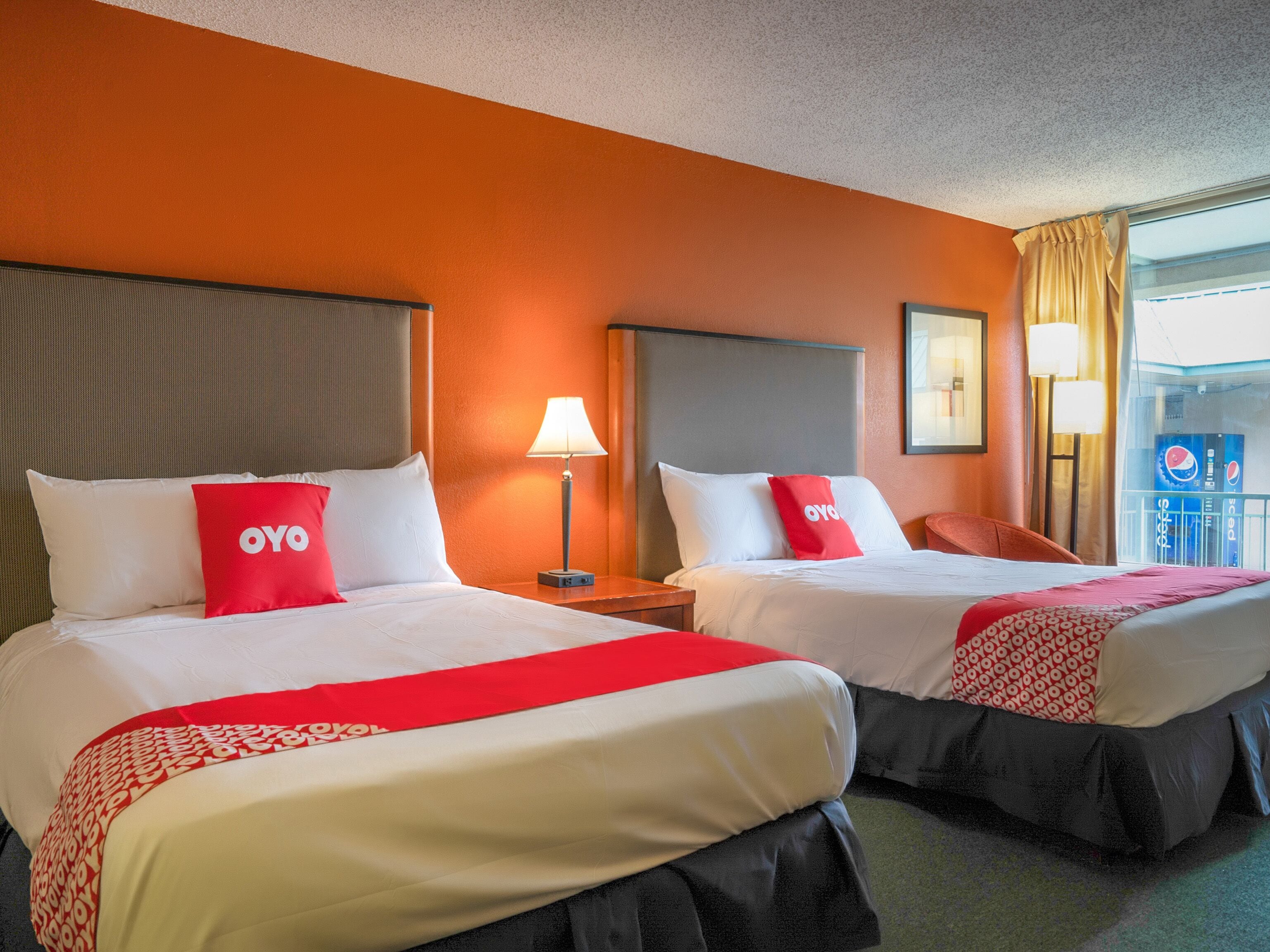 Two double beds with white and red bedding, a red OYO pillow, a grey headboard, a bedside table with a lamp, and a window with curtains.