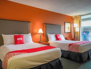Two double beds with white and red bedding, a red OYO pillow, a grey headboard, a bedside table with a lamp, and a window with curtains.