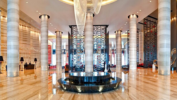 Un moderno vestíbulo con techos altos, grandes columnas cilíndricas, una fuente de agua reflectante en el centro y un mostrador de recepción.