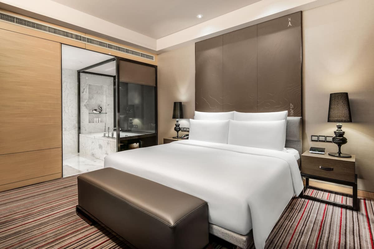 Ein modernes Hotelzimmer mit einem großen Bett, einem Holzschrank, einem Marmorbad und einem roten Teppich mit Streifen.