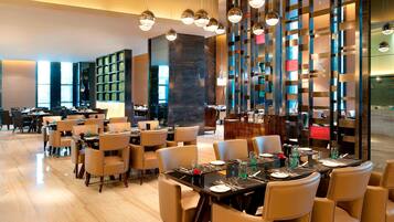 A modern restaurant with multiple dining tables and chairs, a large bookshelf, and a decorative wall with hanging lights.