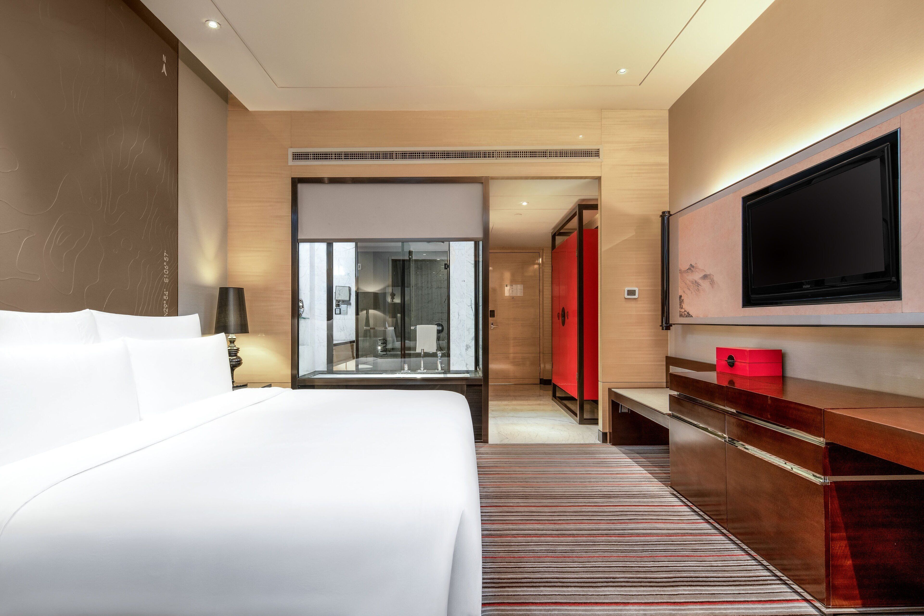 A modern hotel room with a large bed, a flat-screen TV mounted on the wall, a wooden desk with drawers, and a red accent wall.