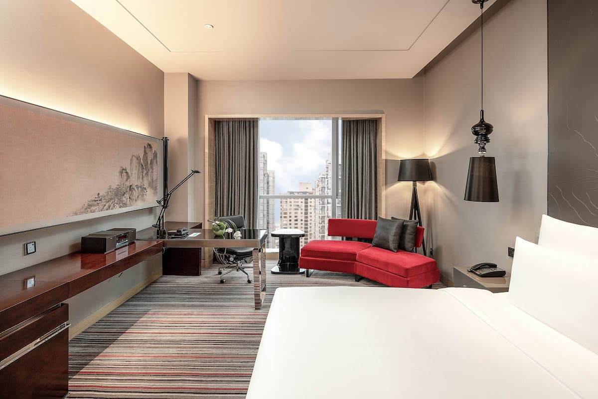 Ein Hotelzimmer mit einem Bett, einem roten Sofa, einem Schreibtisch mit Stuhl, einer Lampe und einem großen Fenster mit Vorhängen.