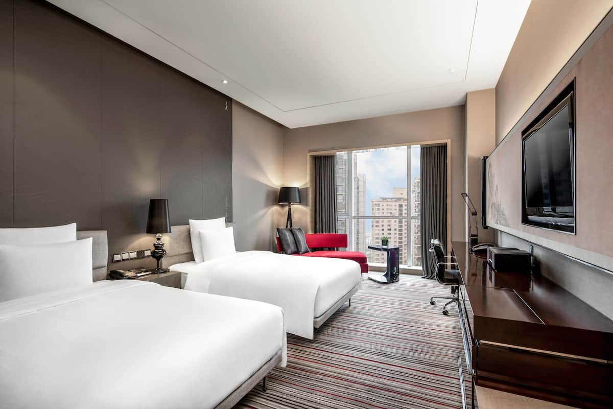 Ein Hotelzimmer mit zwei Betten, einem Schreibtisch, einem Fernseher und einem großen Fenster mit Blick auf die Stadt.