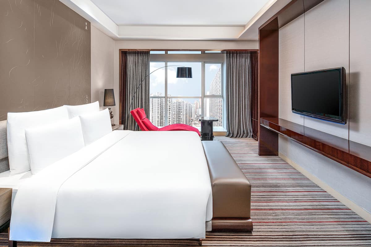 Ein Hotelzimmer mit einem großen Bett, einem an der Wand befestigten Fernseher, einem roten Sessel und Blick auf hohe Gebäude durch das Fenster.