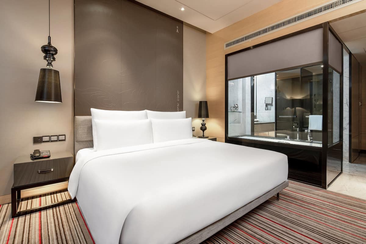 Ein modernes Hotelzimmer mit einem großen Bett, einem Nachttisch mit Lampe, einem zusätzlichen Tisch und einem ein Glas umschlossenen Badezimmer.