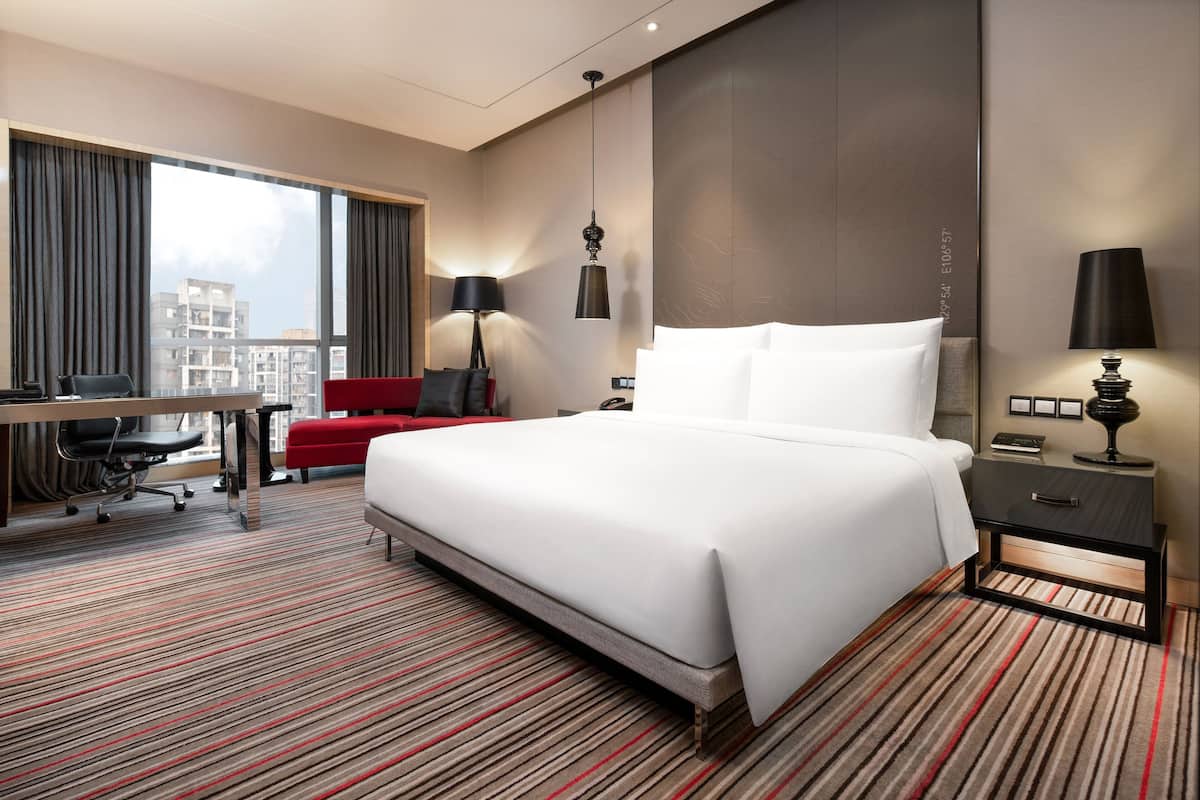 Ein Hotelzimmer mit einem großen Bett, einem Schreibtisch mit Stuhl, einer roten Couch und Blick auf Gebäude durch das Fenster.