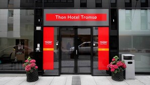 The entrance to Thon Hotel Tromsø with a modern design, featuring a prominent red sign and two potted plants with pink flowers. - Thon Hotel Tromsø (Tromsø)