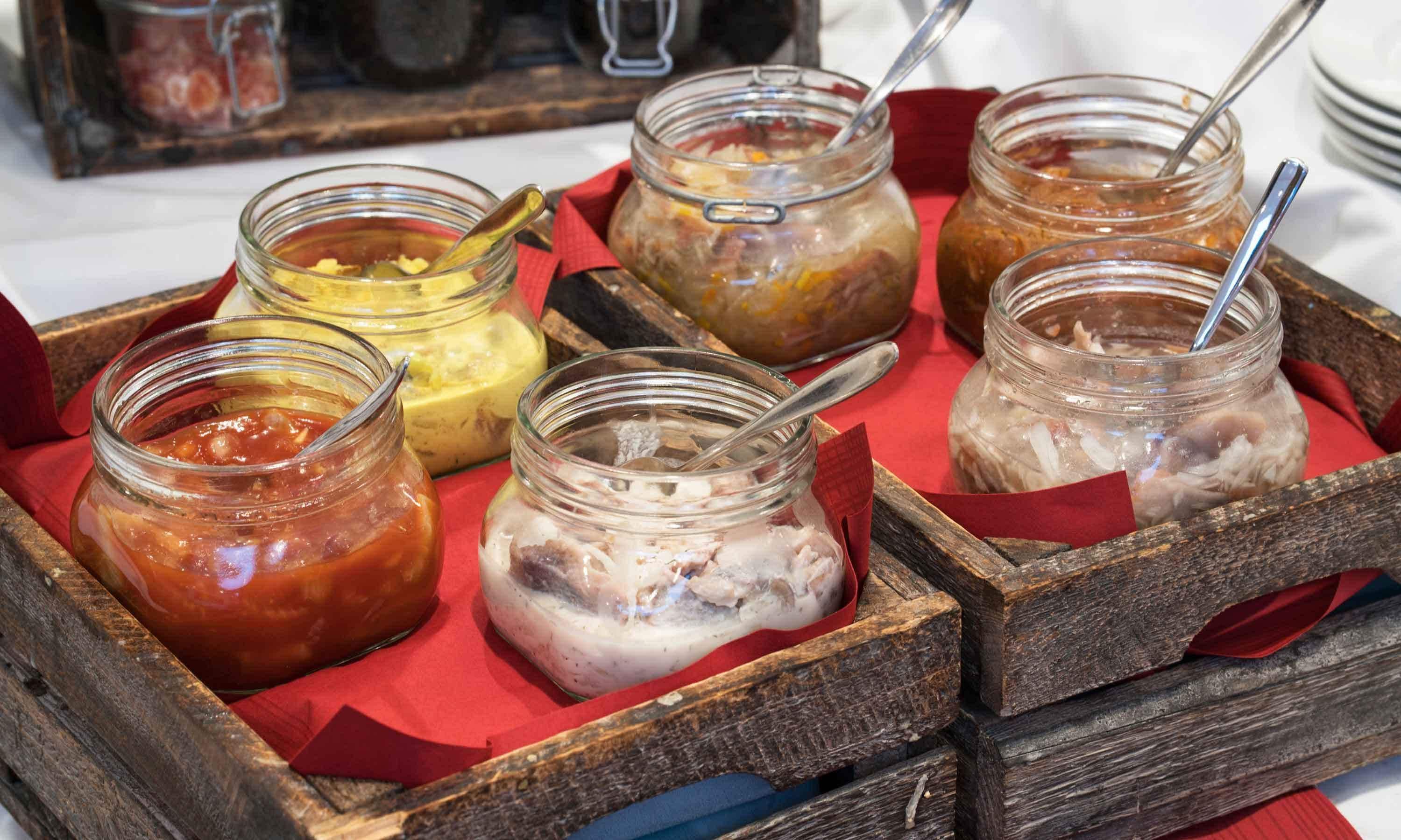 A wooden tray with six glass jars containing different food items, each with a metal spoon.