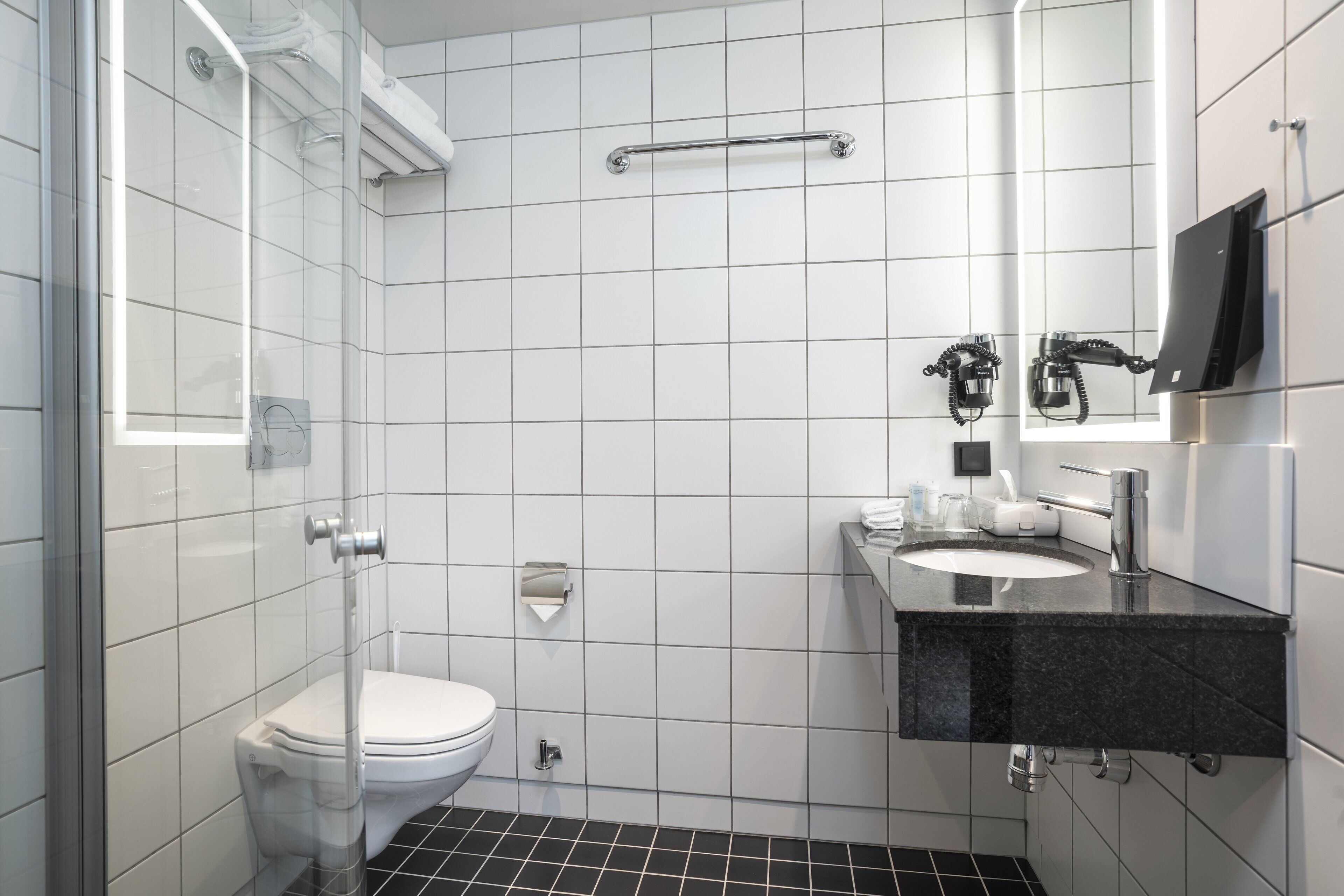 Un bany modern amb una cabina de dutxa de vidre, un vàter blanc, un lavabo negre amb mirall i un suport per a tovalloles.