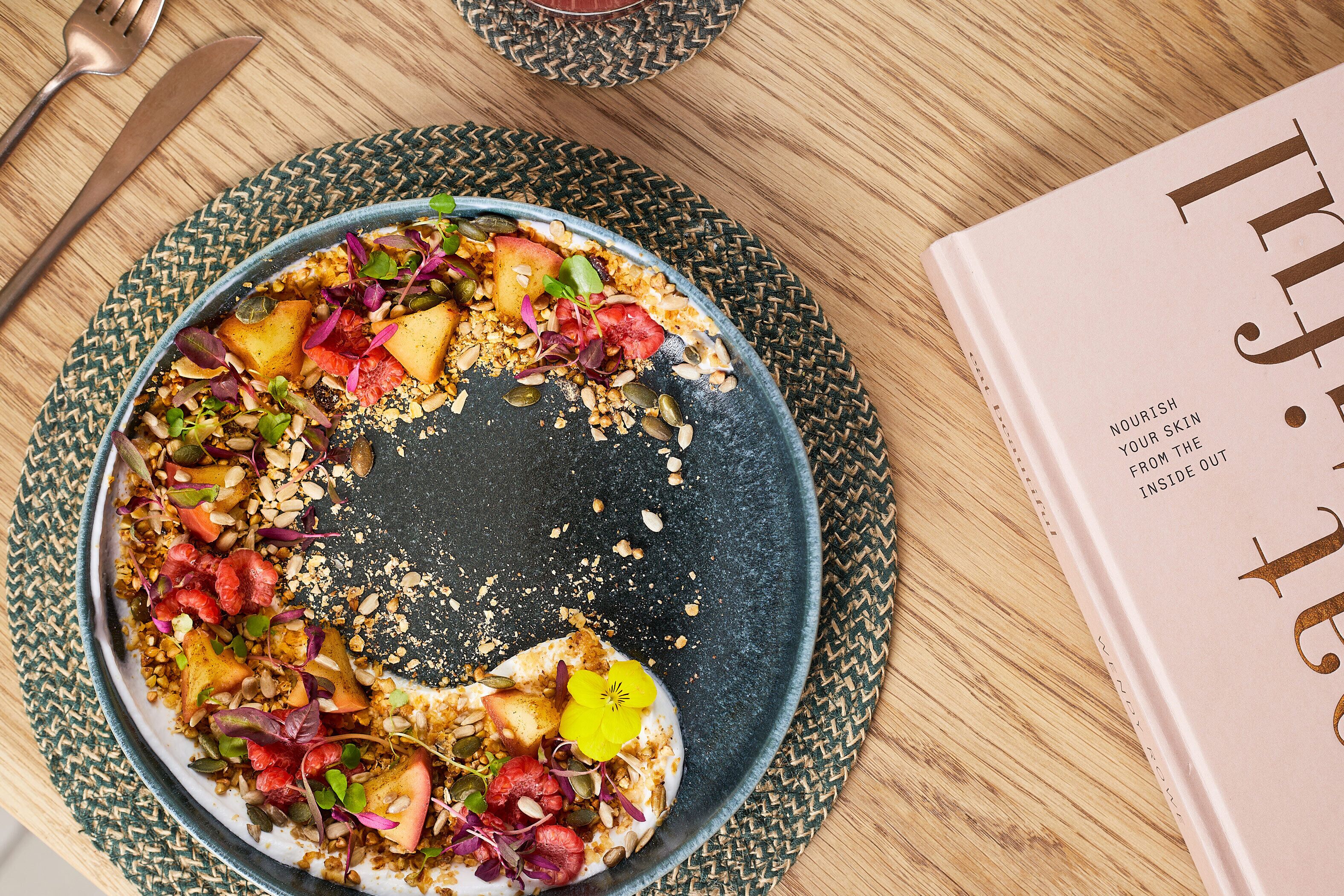 A plate of food with various fruits and seeds on a woven placemat, accompanied by a book titled "Nourish Your Skin From The Inside Out".