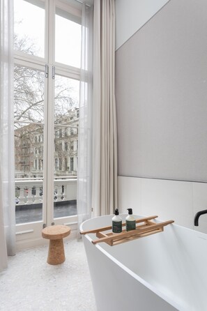 A bathroom with a bathtub, a wooden stool, and a window with a view of buildings. - Inhabit Queen's Gardens, a Member of Design Hotels (London)
