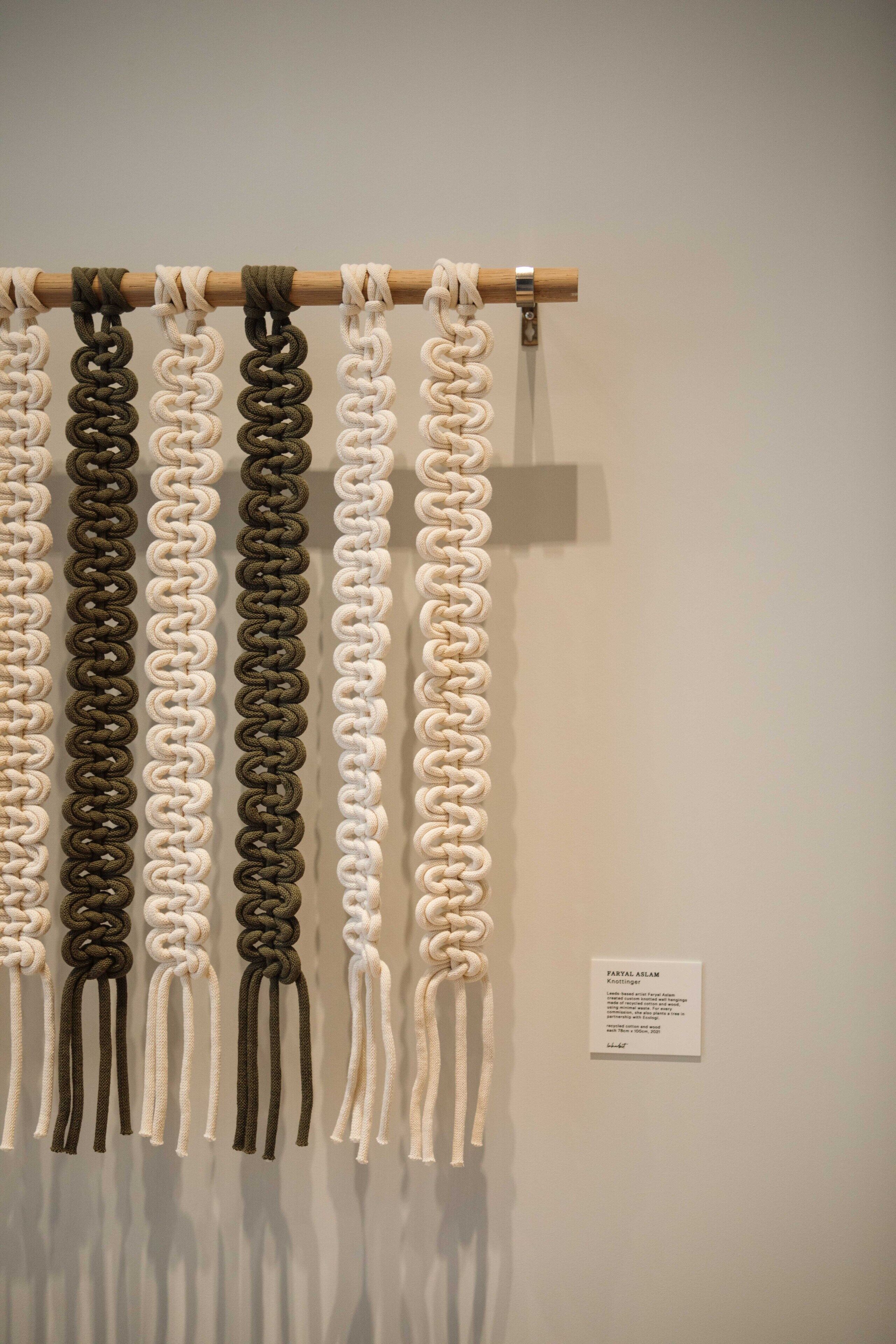 A display of intricately braided cords hanging from a wooden rod, with a label indicating 'FARTAL ASLAM KNOTTING'.