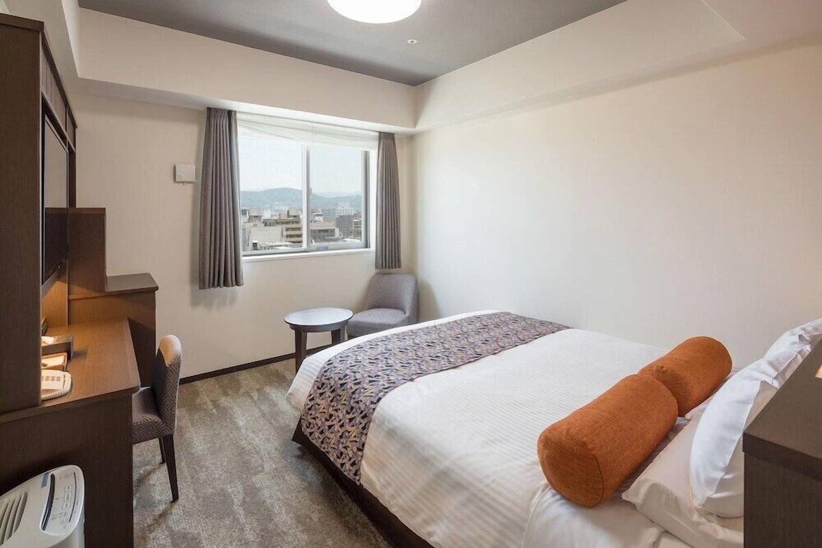 A hotel room with a bed, a desk, a chair, a small table, and a window with a view of the city.