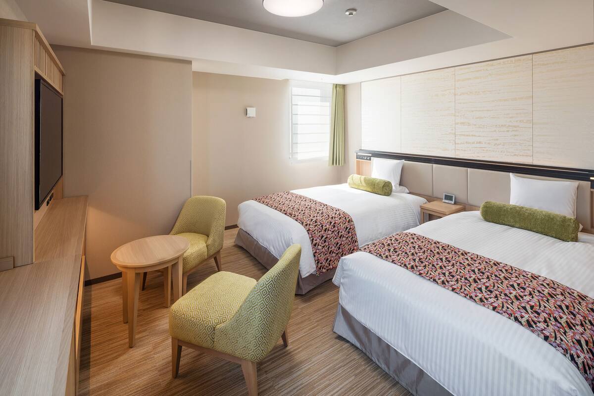 A hotel room with two beds, a small table, two chairs, a TV, and a window with curtains.