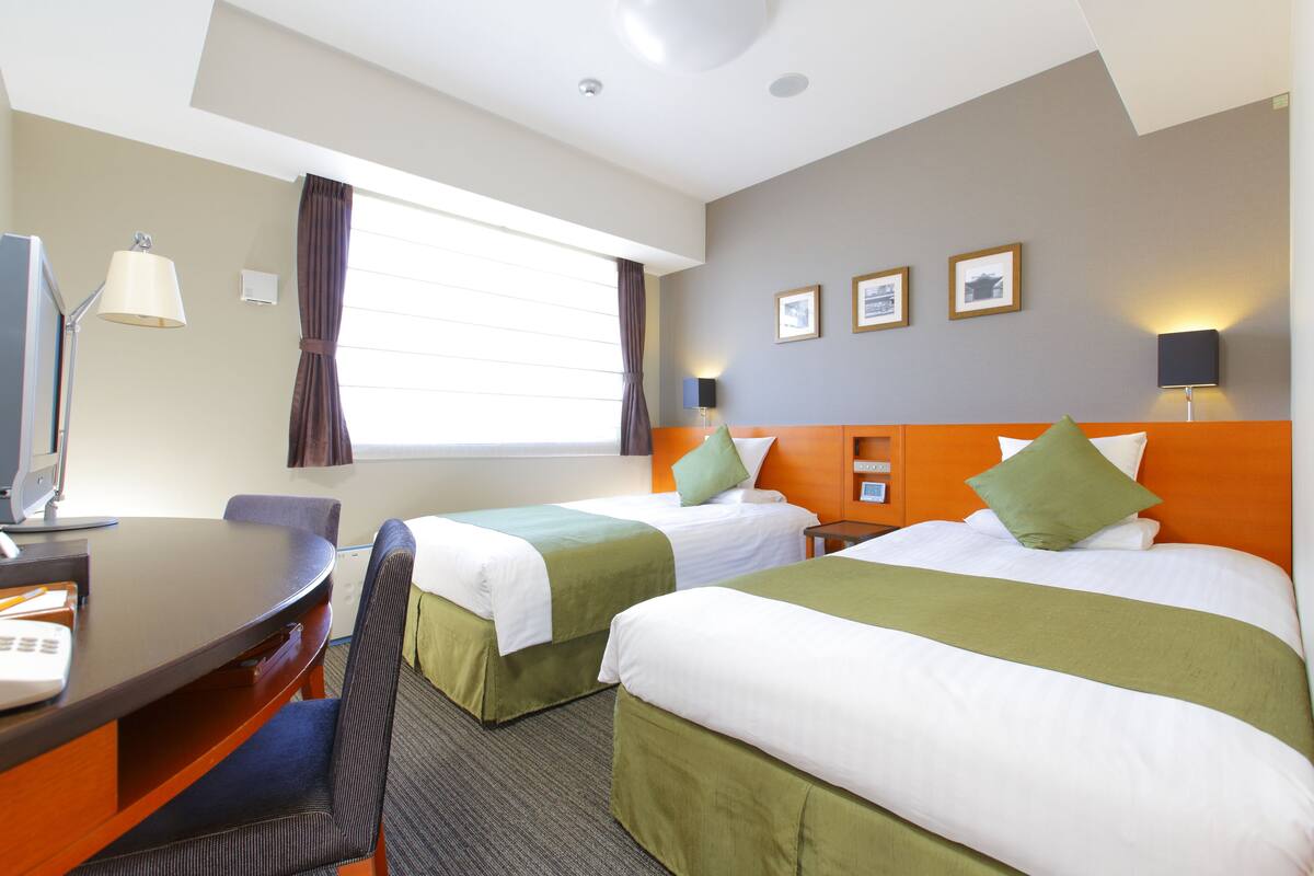 A hotel room with two beds, a desk, and a chair. There are curtains, a lamp, and framed pictures on the wall.