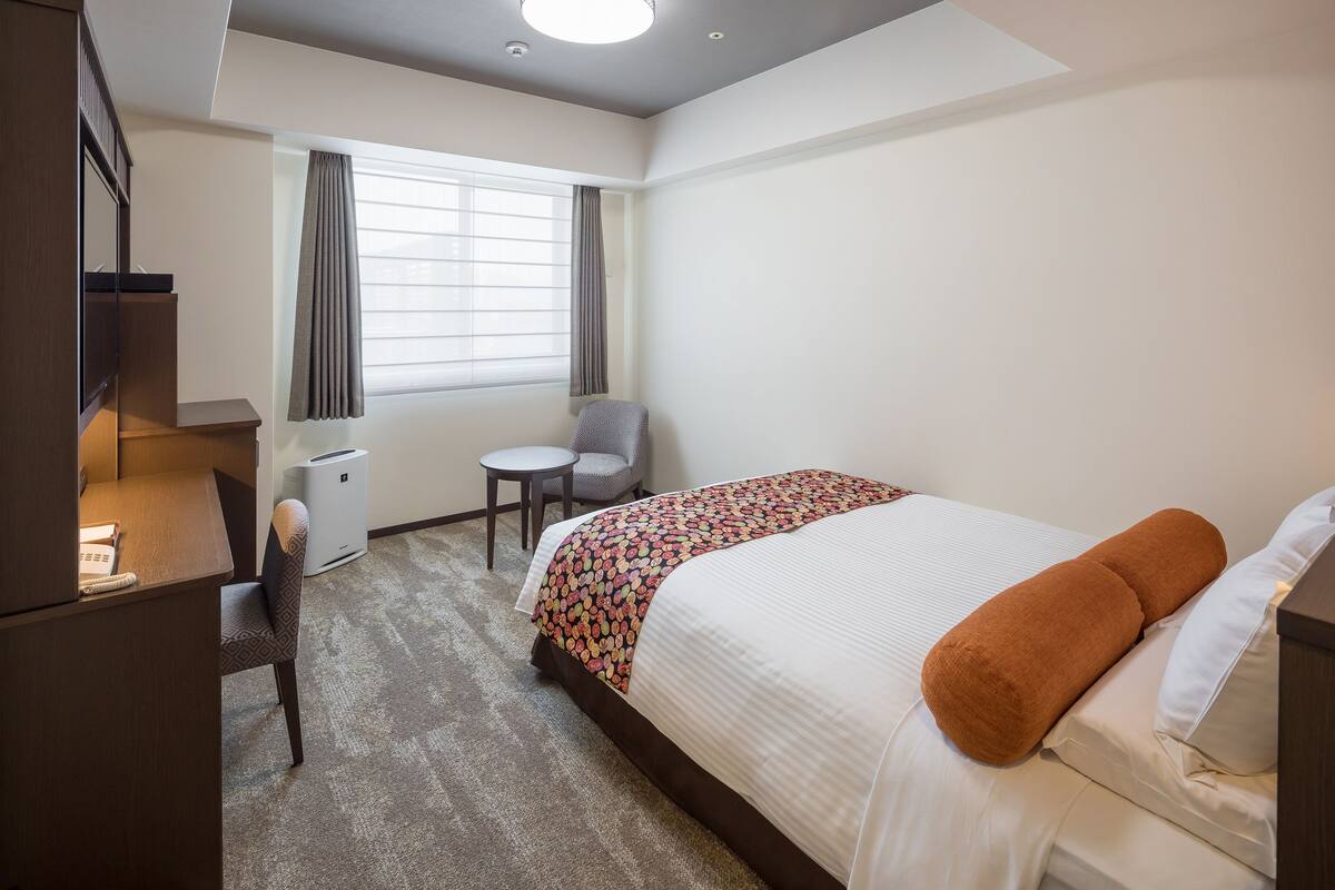 A hotel room with a bed, a desk, a chair, a small table, and a window with curtains.