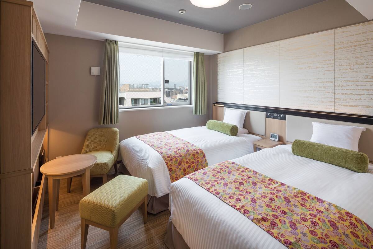 A hotel room with two beds, a small table, a chair, a TV, and a window with a view of the city.