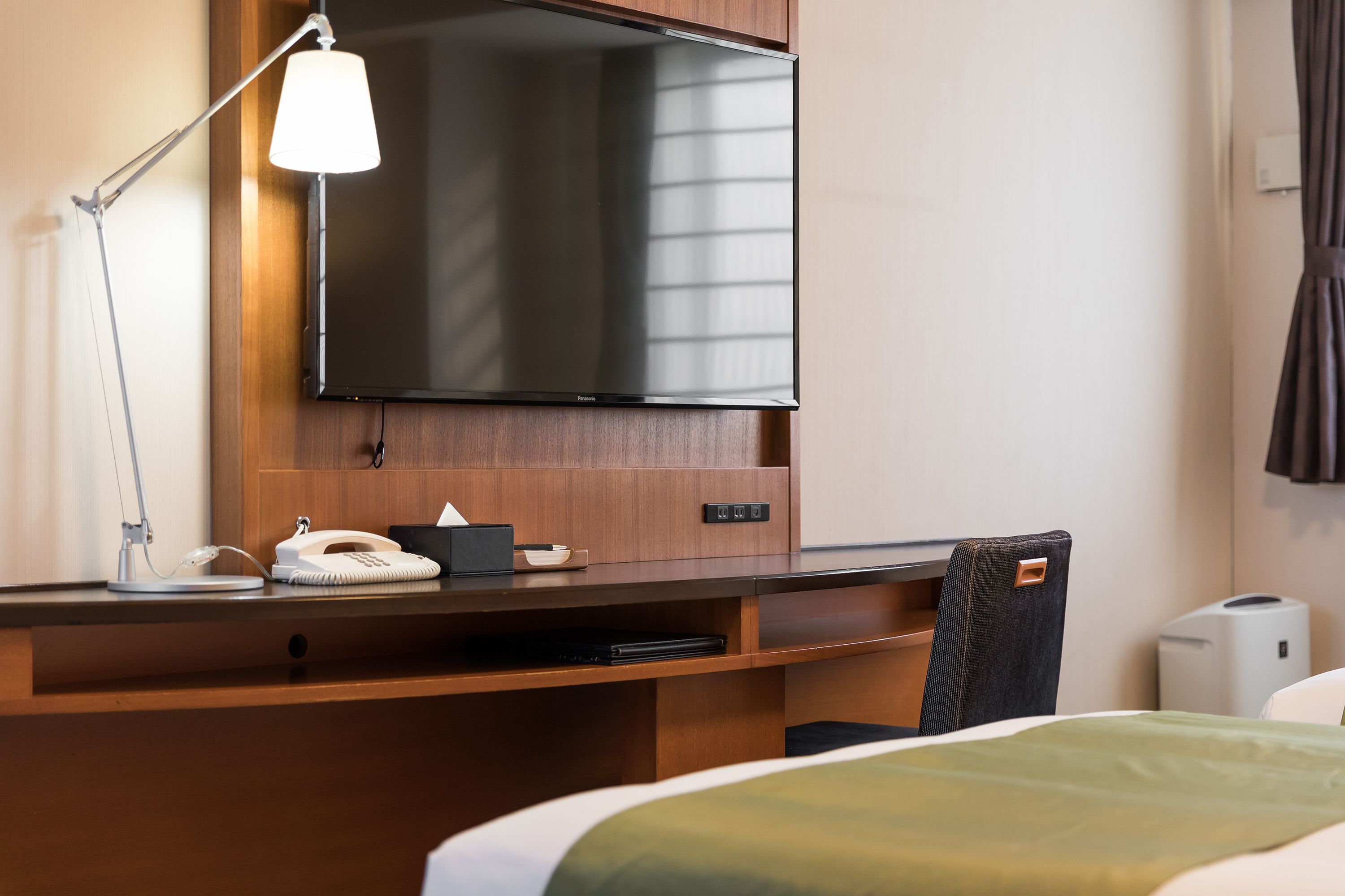 A hotel room with a bed, a desk with a lamp, a telephone, a flat-screen TV, and a minibar.
