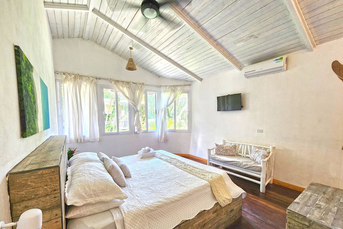A bedroom with a wooden bed, a white sofa, a TV mounted on the wall, and a window with curtains.