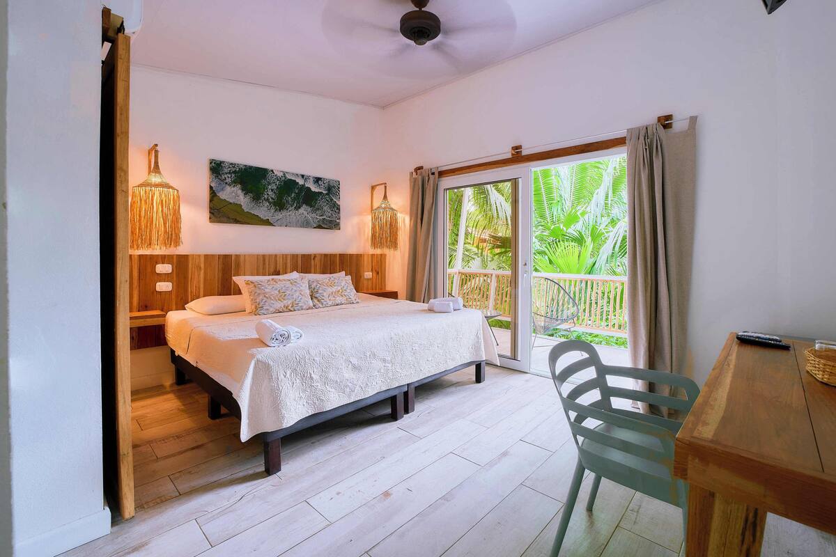 A bedroom with a bed, bedside lamps, a wooden headboard, a window with curtains, and a view of greenery outside.