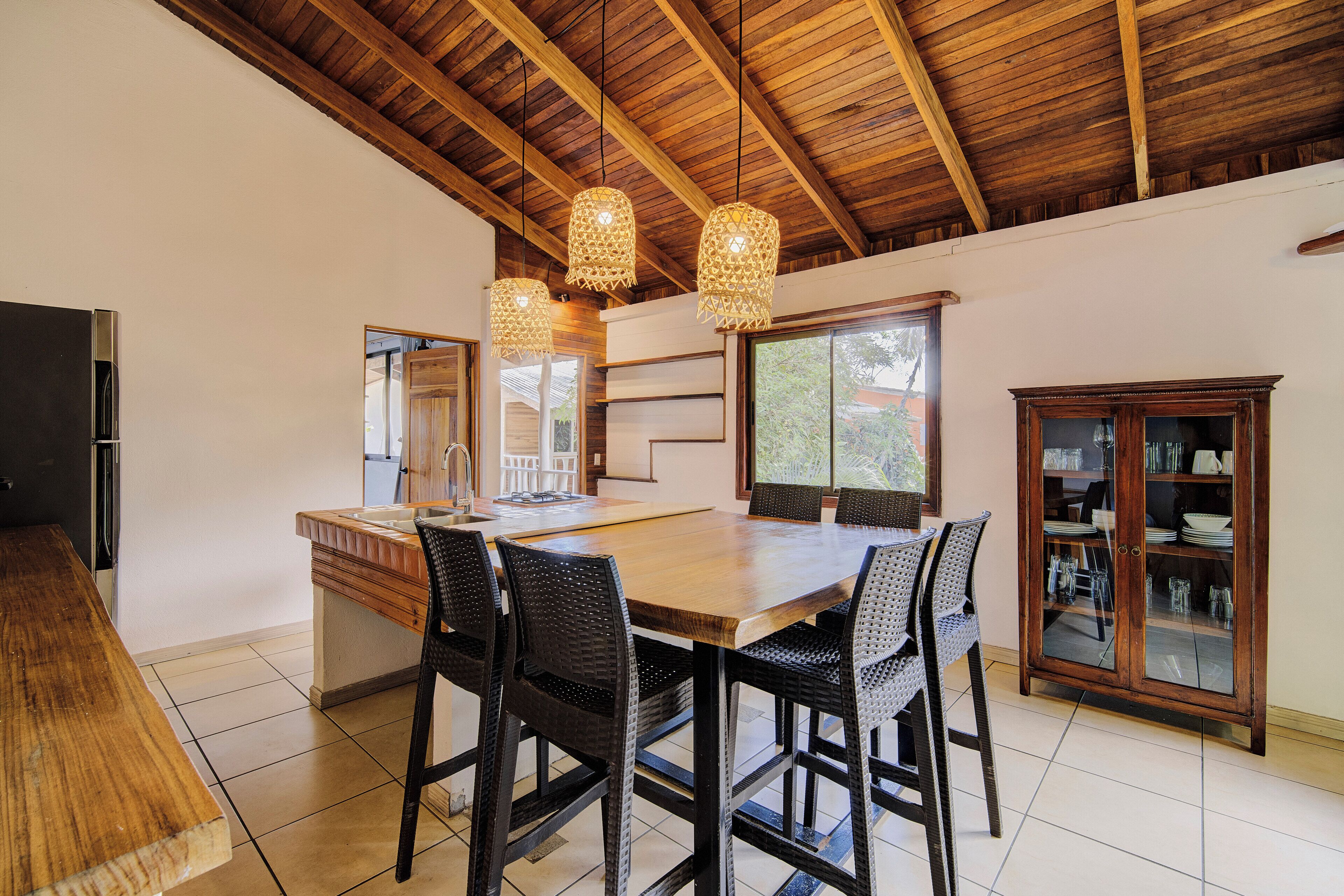 A spacious kitchen with a wooden ceiling, a large island with chairs, hanging lights, and a cabinet with glass doors.