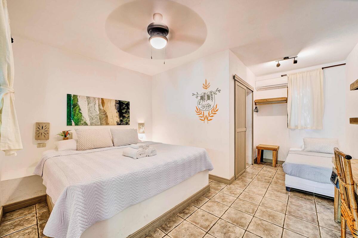 A bedroom with a large bed, a ceiling fan, a wall-mounted air conditioner, and a small bedside table.
