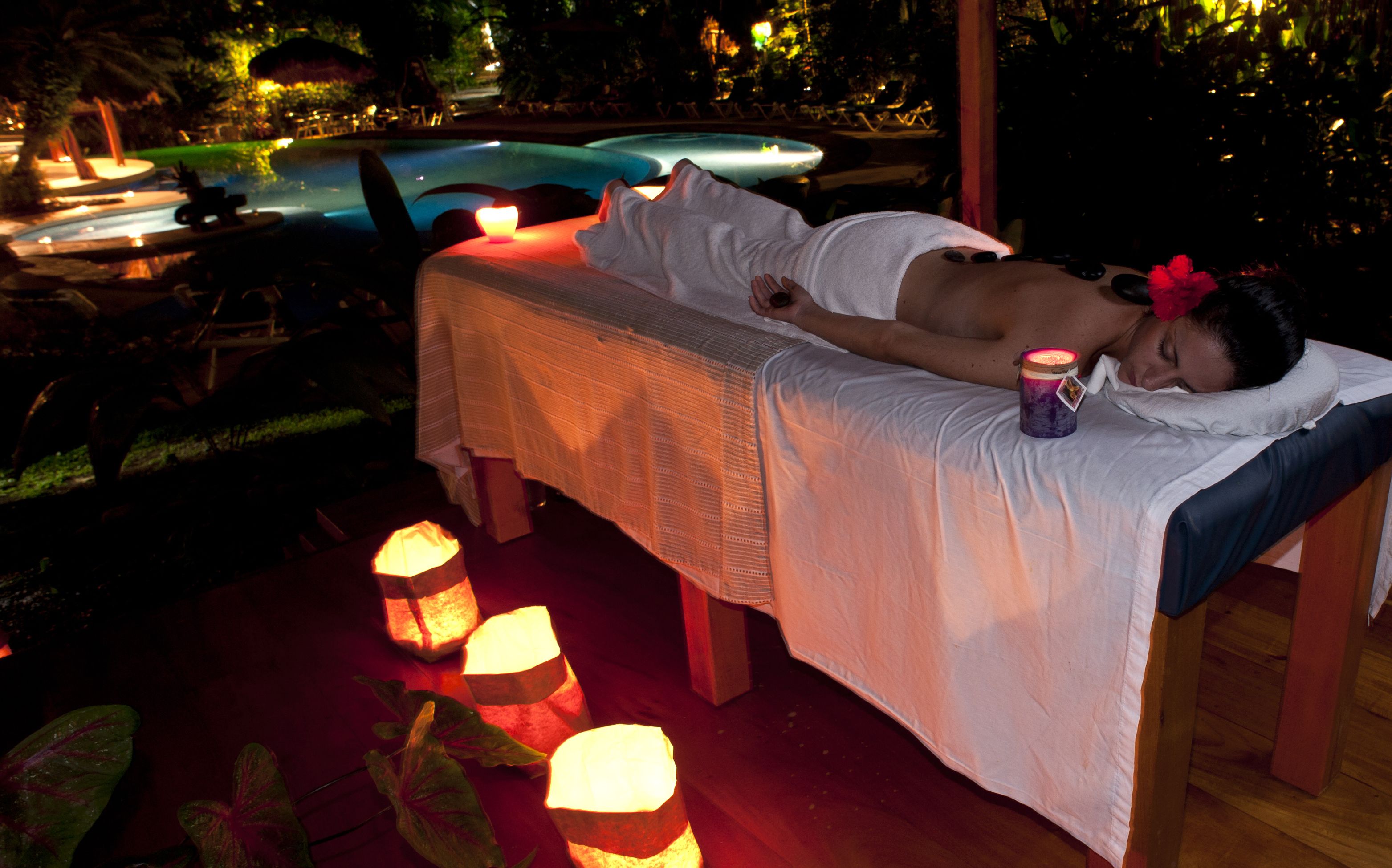 A person lying on a massage table outdoors with candles and a pool in the background.