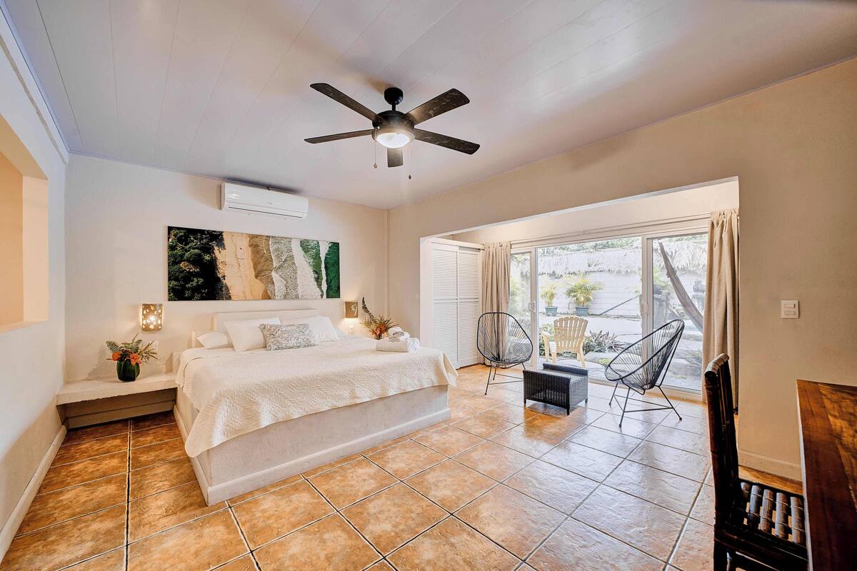 A bedroom with a bed, a ceiling fan, a window with curtains, a chair, and a small table.
