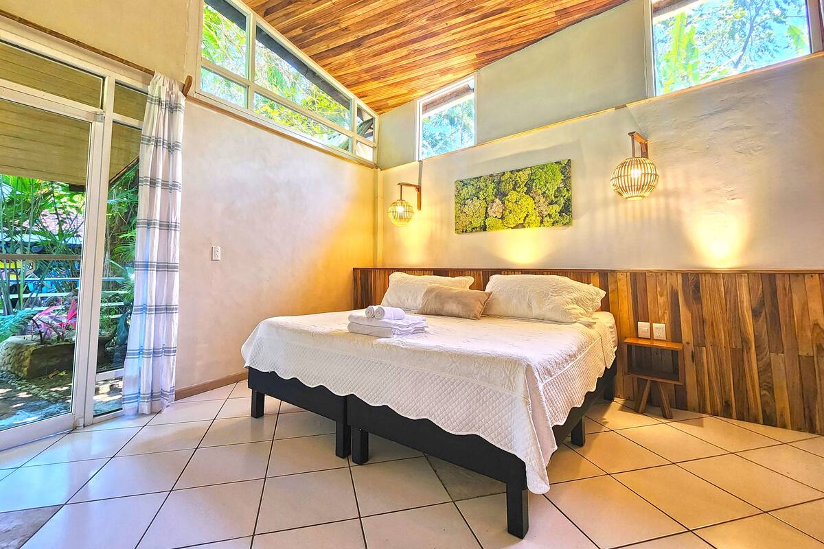 A bedroom with a bed, wooden ceiling, large windows, and a view of greenery.