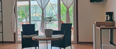 A room with a round table, two chairs, a potted plant, a TV mounted on the wall, and a coffee machine on a shelf.