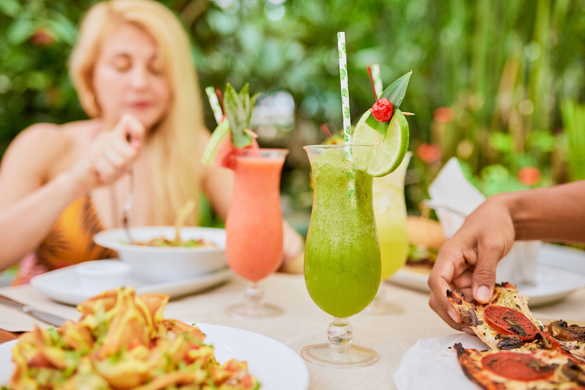 A dining table with various dishes and drinks, including a green cocktail with lime and a red drink with a straw.