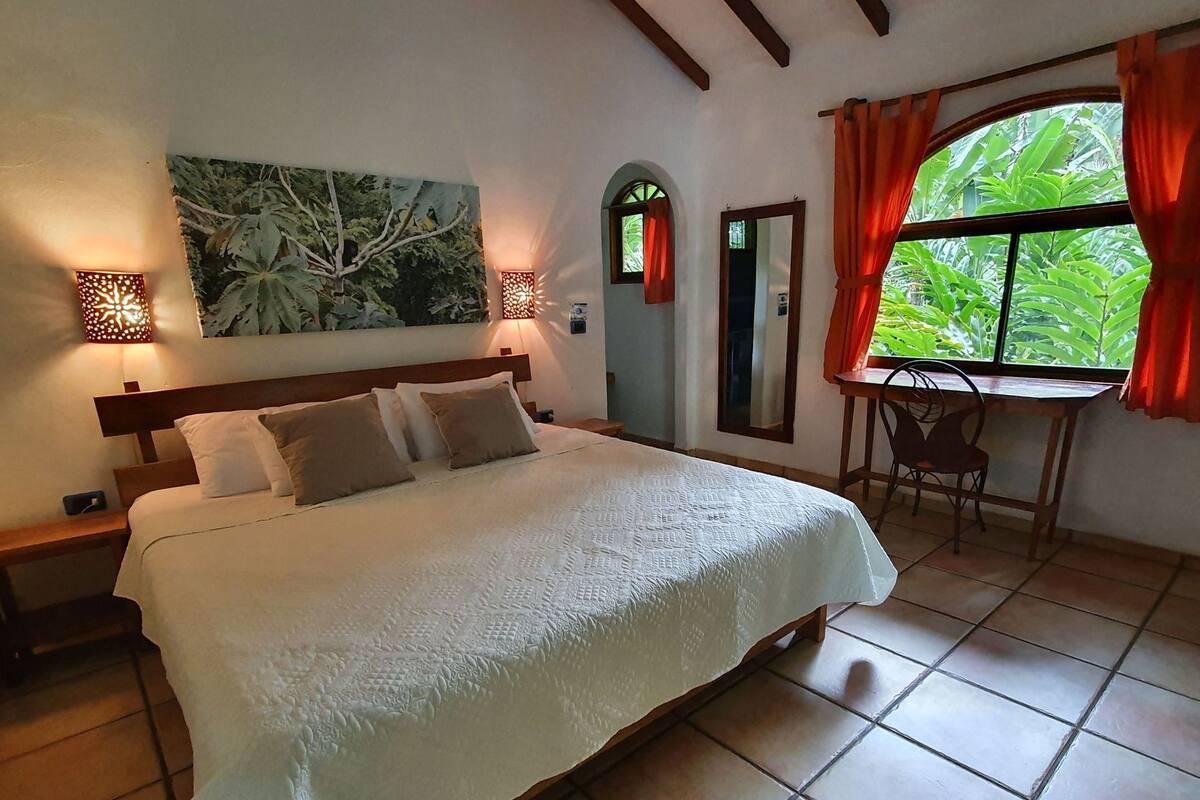 A bedroom with a bed, two bedside tables, a desk with a chair, a window with curtains, and a wall-mounted mirror.
