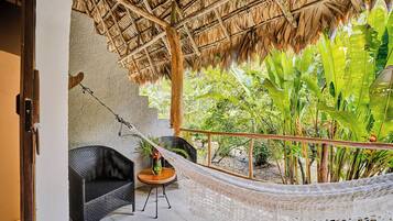 Una hamaca, una silla de mimbre y una planta en maceta en un patio con techo de paja y abundante vegetación.